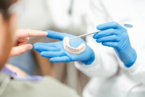 A dentist in blue gloves holds a dental mold and points at it with tweezers as a patient gestures toward the mold, indicating a discussion about dental treatment or procedures.