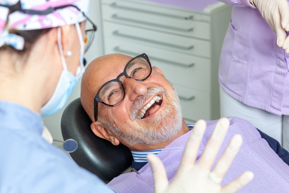 An older man with glasses smiles and laughs while sitting in a dental chair. He wears a bib, while dental staff in masks and gloves stand nearby in a brightly lit clinic room.