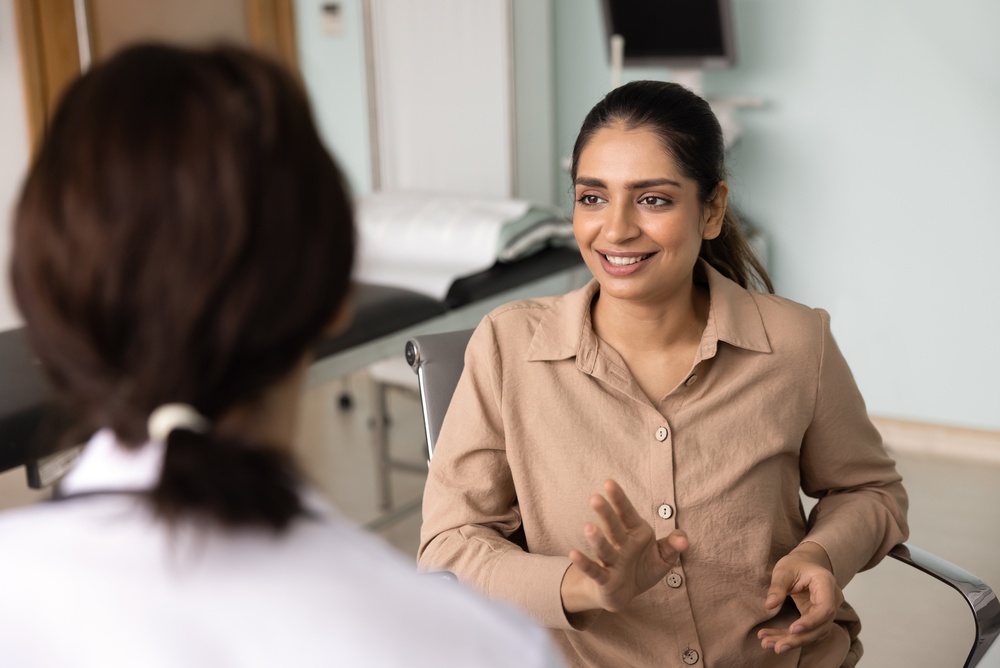 A woman sits in a medical office, smiling and gesturing with her hands while talking to a doctor seen from behind in a white coat. The environment looks friendly and supportive.