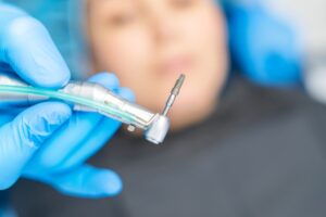 A gloved hand holds a dental drill with an implant attached. In the blurred background, a partially visible patient lies down, wearing a dental bib and cap, getting ready for a dental implant procedure.