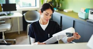 A woman in dark scrubs sits at a desk in a modern office, reviewing paperwork. The background features a chair, desk, plants, office equipment, and natural light streaming through a window.