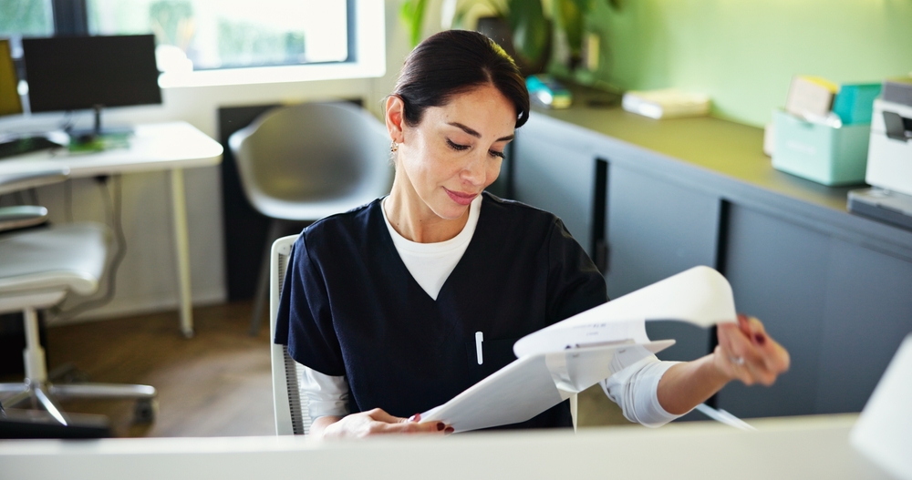 A woman in dark scrubs sits at a desk in a modern office, reviewing paperwork. The background features a chair, desk, plants, office equipment, and natural light streaming through a window.