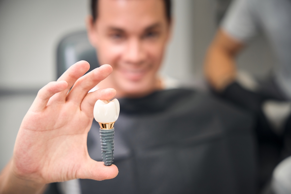 A smiling person in a dental bib holds up a dental implant with a visible artificial tooth, showing it to the camera. The blurred background keeps focus on the implant in their hand.