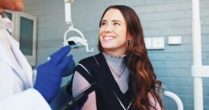 A young woman with long brown hair smiles in a dental chair. A person in a white coat and blue gloves, likely a dentist, speaks to her while holding a dental tool. The background features a light blue-gray brick wall.